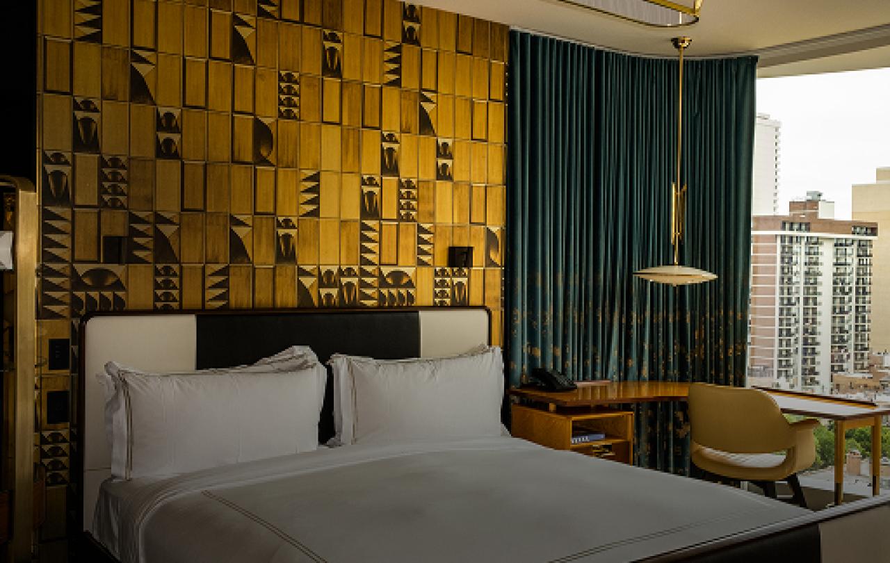 A room with a bed in white linen, gold decor on the wall, and dark blue curtains near a desk at a window.