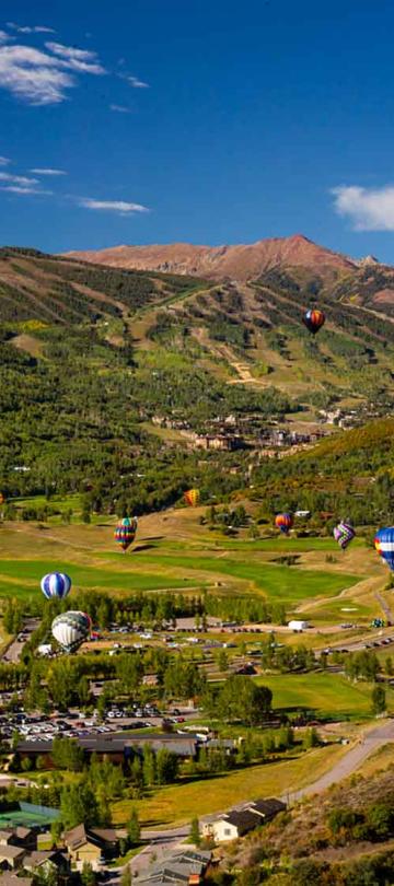 A dozen hot air balloons fly over the town of Snowmass, with the green mountains in the background