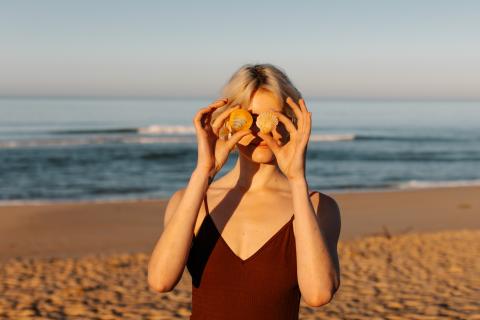 A woman holds sea shells in front of her eyes like sunglasses, as she stands with her back to the sea.