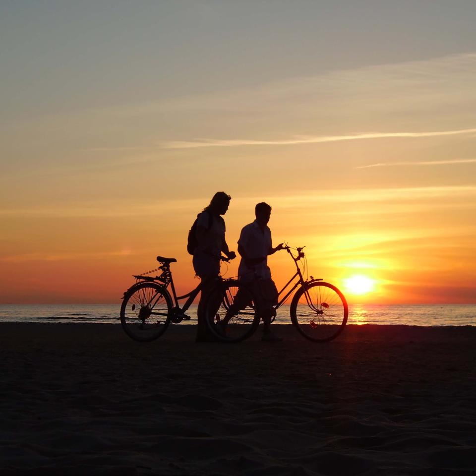 shadows of two people walking beside bikes on the beach at sunset