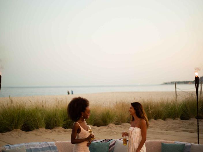 two women stand near a firepit surrounded by coaches on the beach
